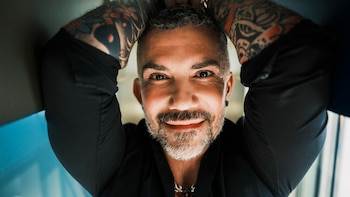 Primer plano de Tristan Pineiro, hombre con cabello gris corto y barba, sonriendo. Tiene brazos levantados detrás de la cabeza y tatuajes coloridos en ambos brazos
