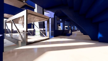 Interior moderno con estructuras arquitectónicas en azul oscuro y piso claro. Destaca una construcción de metal y vidrio, con luz solar que proyecta sombras