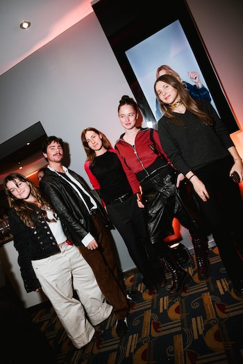 Un grupo de cinco jóvenes, tres mujeres y dos hombres, posan de pie en un interior con luz tenue y un patrón en el suelo, junto a una pantalla vertical
