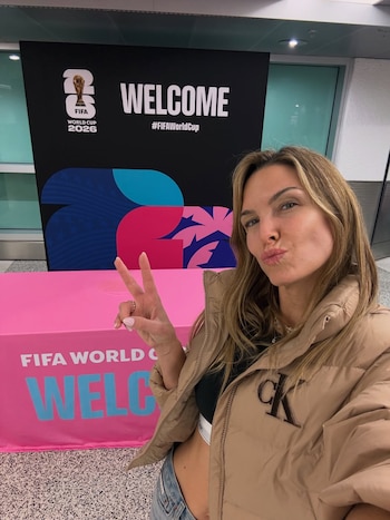 Mujer posa con los labios fruncidos y la mano haciendo el signo de la paz, frente a un cartel negro de la Copa Mundial FIFA 2026 y un mantel rosa