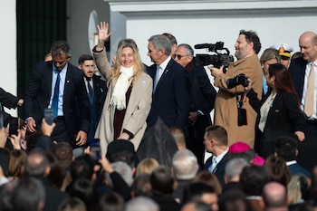 Una mujer rubia con abrigo beige y bufanda blanca sonríe y saluda con la mano, rodeada de hombres de traje y una multitud que sostiene teléfonos para fotografiar