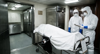 Dos personas con trajes de protección personal, visores y mascarillas, junto a una camilla con un cuerpo cubierto por una sábana blanca en una morgue
