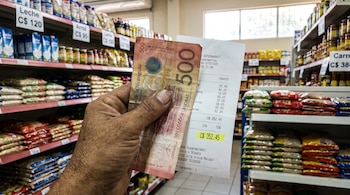 Mano sostiene billete de 500 córdobas y factura de supermercado con 15% de IVA; de fondo, estanterías con productos y etiquetas de precios.