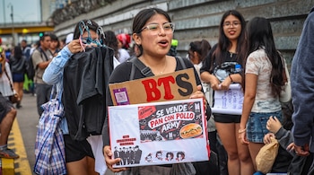 Una mujer con gafas sostiene un cartel de cartón con la palabra BTS y un letrero impreso en una calle concurrida.