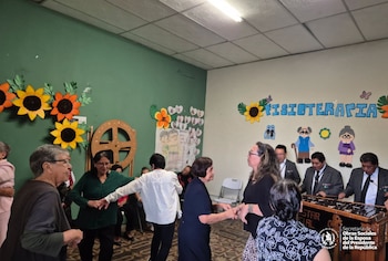 Varias personas mayores bailan en un salón decorado con girasoles de papel, mientras tres hombres tocan la marimba al fondo