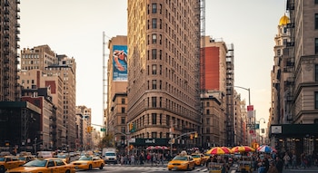 Vista da rua de Nova York com o Flatiron Building triangular no centro, cercado por táxis amarelos, pedestres e arranha-céus sob um céu claro.