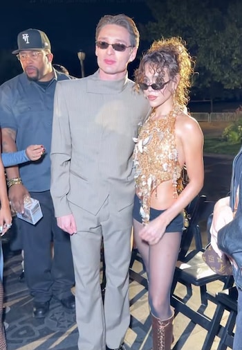 Un hombre con traje claro y gafas de sol posa junto a una mujer con top dorado ornamentado y gafas oscuras en un espacio exterior de noche