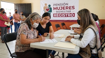 Mujer adulta mayor con cubrebocas escribe en un formulario sobre una mesa, asistida por una mujer joven. Otra mujer con chaleco también manipula papeles.