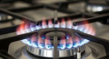 Primer plano de una hornalla de cocina a gas encendida, mostrando una llama azul y anaranjada alrededor de un quemador metálico. Se ven las rejillas de soporte negras.
