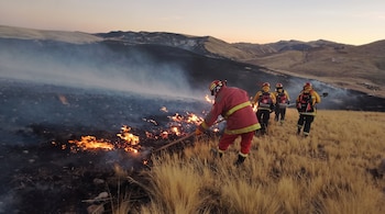 Cuatro bomberos con cascos y trajes de protección apagan el fuego en un campo de pasto seco junto a cerros.