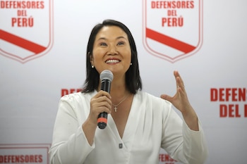 Keiko Fujimori, sonriendo, habla por un micrófono con blusa blanca y collar de cruz, gesticulando, con el logo "Defensores del Perú" al fondo