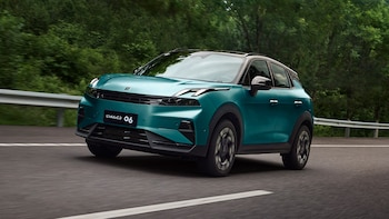 Automóvil Lynk&Co 06 de color verde azulado en movimiento sobre una carretera, con árboles verdes borrosos al fondo y una barandilla lateral
