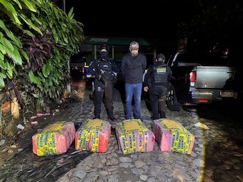 Un hombre custodiado por dos agentes policiales junto a cuatro grandes paquetes rectangulares envueltos con cinta amarilla de evidencia en una calle