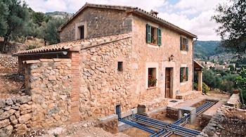 Casa de piedra con tejado de tejas, ventanas con contraventanas verdes y tuberías azules y grises al descubierto en una zanja frontal.