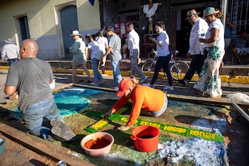 Un hombre con gorra azul crea una alfombra de aserrín colorida en el suelo, con diseños de un sol radiante y dos estructuras arquitectónicas coloniales, en una calle empedrada