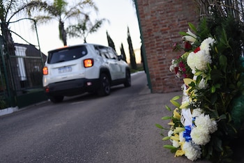 Un SUV blanco sale por un portón de metal verde, con palmeras al fondo y una corona fúnebre de flores blancas, amarillas y rojas en primer plano