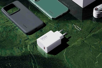 Un cargador blanco de 90 W, un teléfono verde, una funda gris, un cable y una herramienta de extracción de SIM sobre una superficie verde