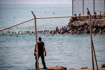 Hombre de espaldas en primer plano; mar; grupo de personas nadando; grupo de personas sobre rocas; valla metálica; varios agentes de seguridad