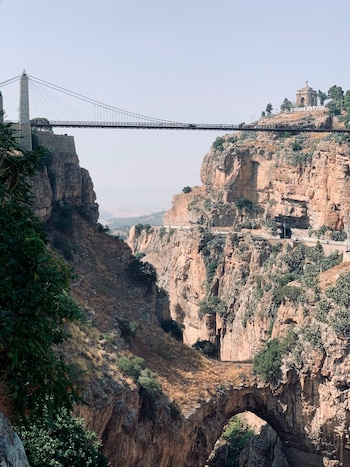 Puente colgante entre dos acantilados de piedra sobre un cañón con vegetación, un arco de roca natural en la base y un monumento al fondo