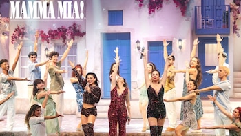 Título Mamma Mia! en la parte superior. Grupo de actores con brazos levantados en escenario, vestuario variado, frente a telón de fondo de casas blancas y flores