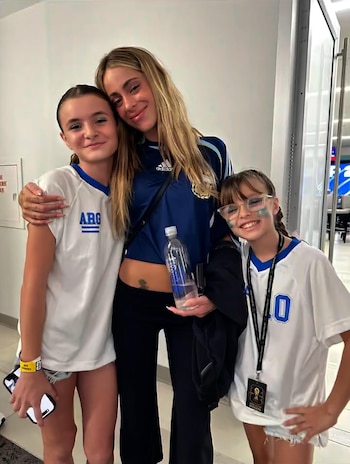 Una mujer rubia y dos niñas sonríen a cámara. La mujer viste una camiseta deportiva azul de Argentina y las niñas, camisetas blancas con números y letras azules