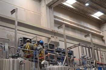 Vista trasera de dos trabajadores con cascos y arneses de seguridad operando paneles de control eléctricos y tuberías de acero inoxidable en una fábrica