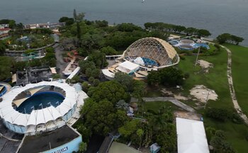 Vista aérea de un complejo de edificios, piscinas, estructuras con techos blancos y una cúpula geodésica, rodeado de árboles, caminos y un cuerpo de agua