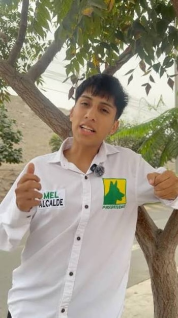 Un hombre joven con camisa blanca y distintivos políticos gesticula frente a la cámara junto al tronco de un árbol