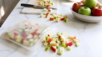 Cubos de manzana verde y roja junto a una bolsa transparente, un recipiente con manzanas enteras y una tabla de cortar con cuchillo.