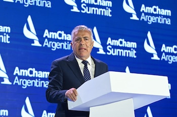 Hombre de cabello gris con traje y corbata hablando en un podio blanco frente a una pantalla azul con logos repetidos de AmCham