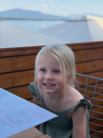Una niña rubia de ojos azules sonríe en primer plano; detrás, una mesa con un menú, una baranda de madera y un paisaje lacustre con montañas