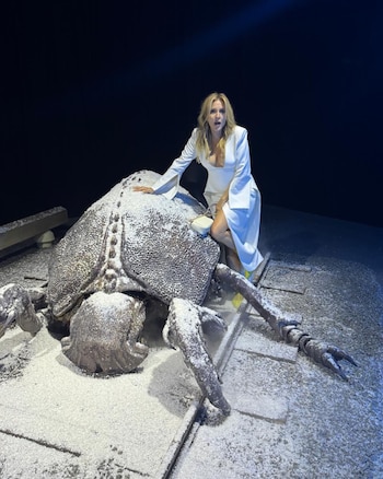 Carla Peterson, con cabello rubio y vestido blanco, posa sentada sobre una gran escultura de un insecto oscuro cubierta de polvo blanco, con fondo oscuro