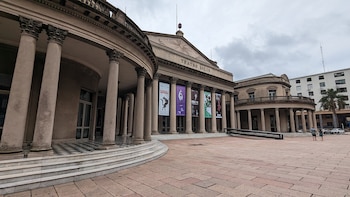 Fachada del Teatro Solís, edificio neoclásico con numerosas columnas, balcones y banderas promocionales, frente a una plaza adoquinada. Cielo gris nublado
