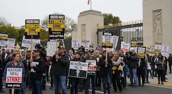 Multitud de manifestantes sosteniendo pancartas sobre huelga, seguro médico y residuals, frente a una gran reja dorada y un edificio beige con una bandera de EE. UU.