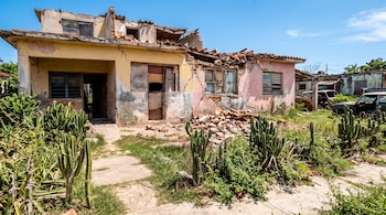 Casa de dos niveles con paredes agrietadas, techo de tejas destruido, escombros en el suelo y vegetación en el patio.
