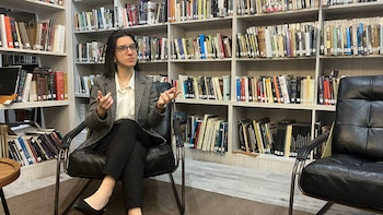 Mujer con gafas y blazer gris sentada en un sillón negro, gesticulando con las manos frente a estanterías de madera repletas de libros en una biblioteca