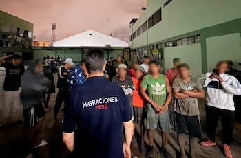 Un agente con camiseta de Migraciones Perú se encuentra de espaldas frente a un grupo de hombres en un patio exterior durante la noche