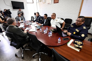 Personas con traje y agentes de policía sentados ante una mesa ovalada con computadoras y botellas de agua