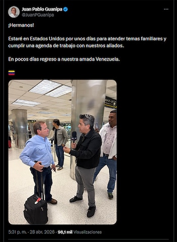 Juan Pablo Guanipa, con camisa azul, sostiene una maleta. Un reportero lo entrevista con micrófono en un aeropuerto, con otras personas de fondo