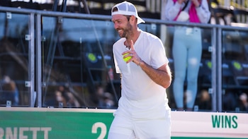 Hombre sonriente con gorra blanca y ropa deportiva blanca sostiene una pelota de tenis amarilla en la mano derecha en una cancha al aire libre