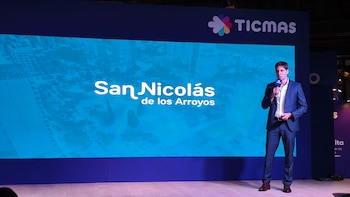 Un hombre de traje azul y camisa blanca habla con un micrófono en un escenario. Detrás, una pantalla azul grande muestra "San Nicolás de los Arroyos" y el logo de TICMAS