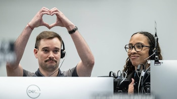 Un hombre y una mujer sentados en escritorios con auriculares. El hombre, con bigote, hace un corazón con las manos sobre su cabeza, mientras la mujer sonríe