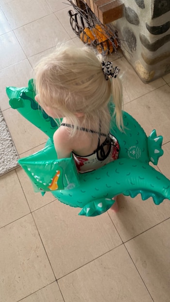 Vista aérea de una niña rubia con coletas y lazo, vista de espaldas, usando un traje de baño floral y un flotador verde de cocodrilo. Está de pie sobre baldosas claras