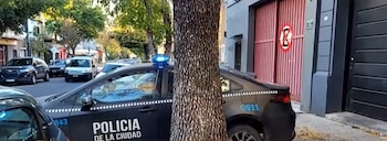 Una patrulla de la Policía de la Ciudad de color oscuro con luces azules en el techo, estacionada en una calle urbana. Un árbol grueso bloquea parcialmente el coche