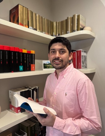 Un hombre con camisa rosada sostiene un libro abierto frente a varias estanterías blancas con libros