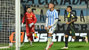 Tres futbolistas en un campo de juego: uno de Racing celebra un gol, el portero y otro jugador de Defensa y Justicia están cerca del arco con un balón