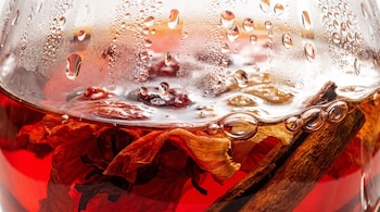 Un recipiente de vidrio contiene líquido rojo, trozos de flor de jamaica y palos de canela; gotas de vapor y burbujas se observan en la superficie.