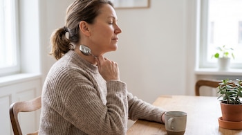Una mujer de perfil con el cabello recogido se masajea el cuello con una cuchara de metal, sentada en una mesa de madera. Hay una taza y una planta.