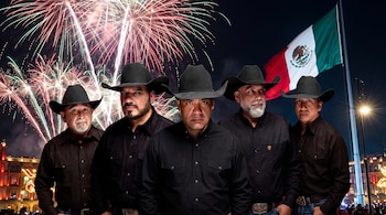 Cinco hombres con sombrero negro y camisa oscura posan frente a fuegos artificiales y una bandera de México ondeando en la noche.
