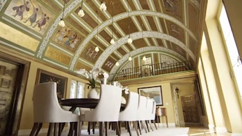 Interior de un gran salón con un techo abovedado ricamente decorado con frescos y detalles dorados, incluyendo un área de comedor con sillas blancas y una mesa